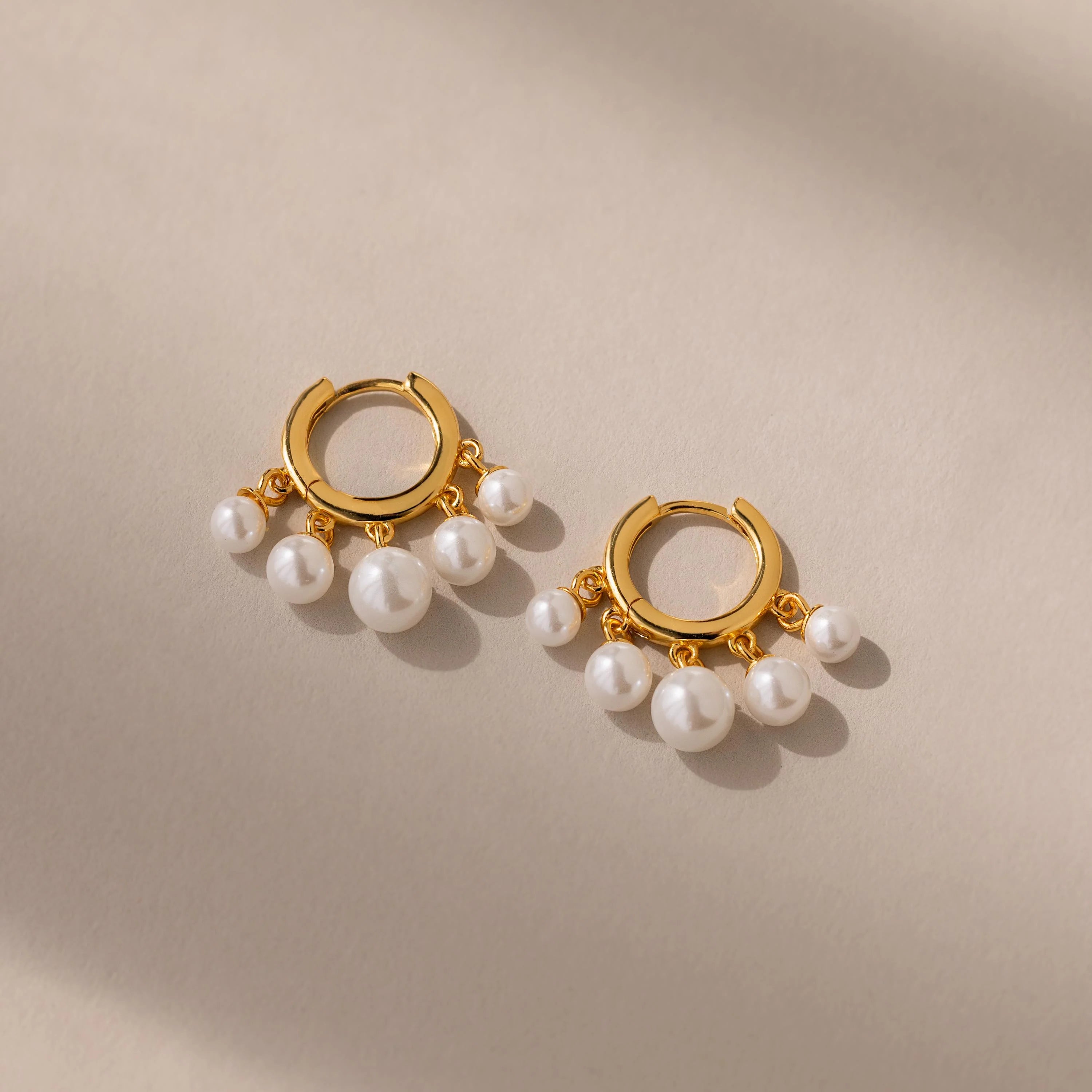 A pair of gold-plated huggie earrings with dangling white pearl beads on a beige background.